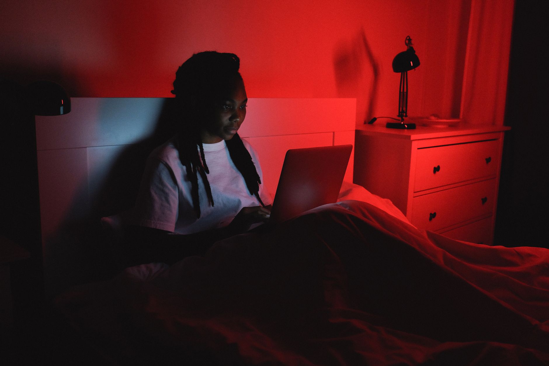 A woman sitting in bed using a laptop, lit by warm ambient light.