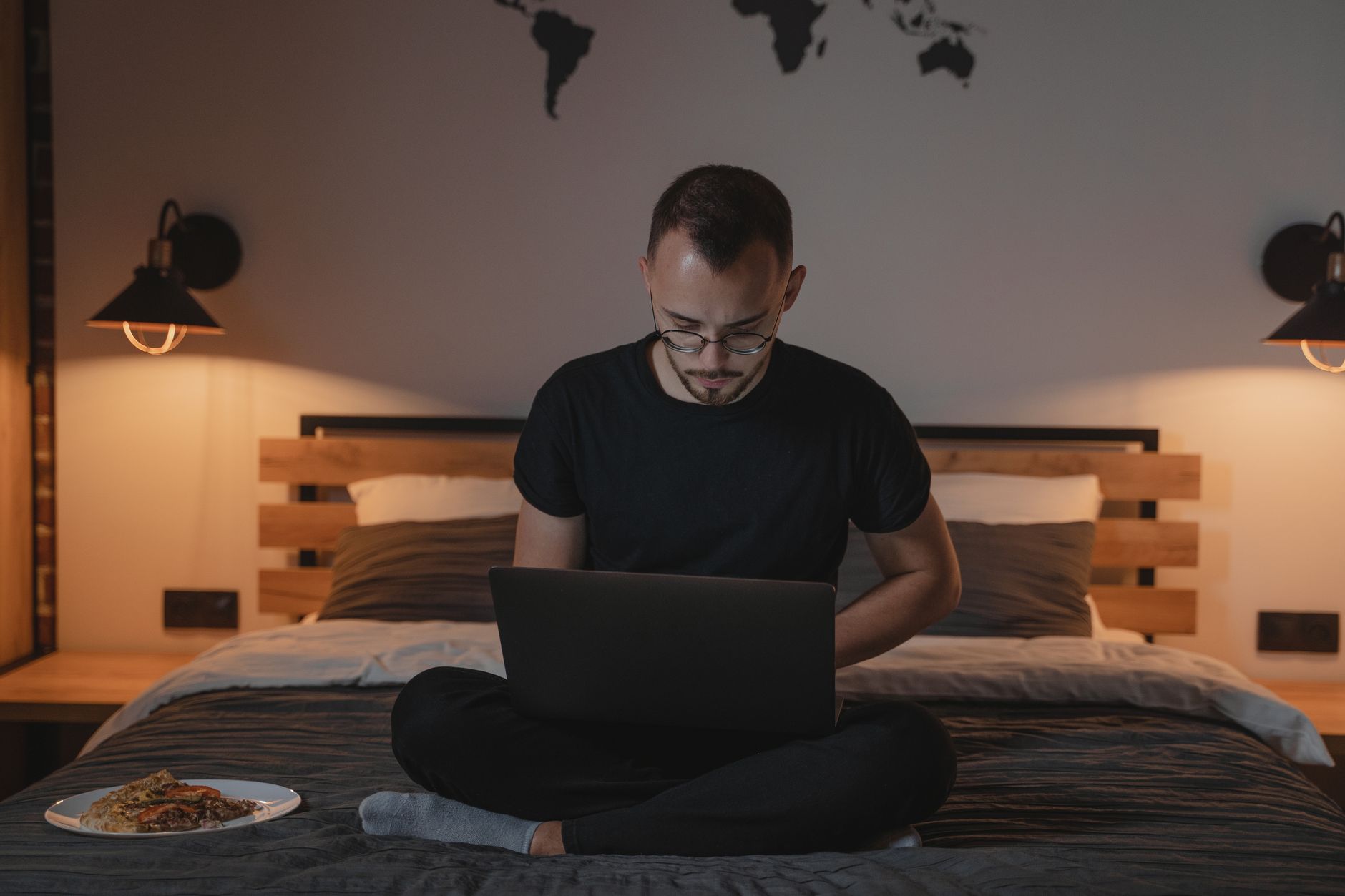 A man using a laptop while sitting on a bed, with warm decor and a wall map in the background.